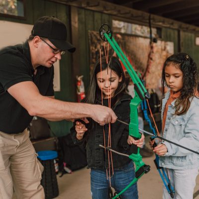 Adult male archery coach teaching adorable little multiethnic girls to shoot with bow and arrow during classes in range