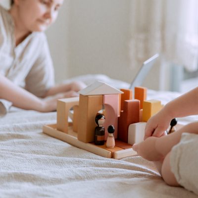 Faceless toddler girl sitting on bed and playing with wooden blocks and toys while mother using laptop