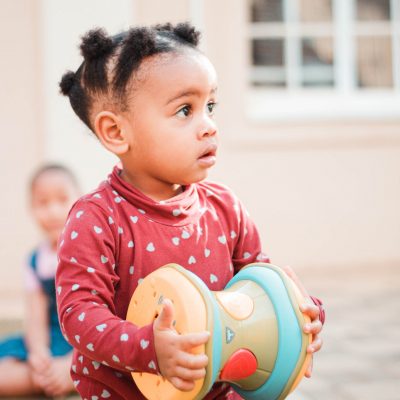 Funny little African American girl with pigtails carrying toy while playing with friend in kindergarten